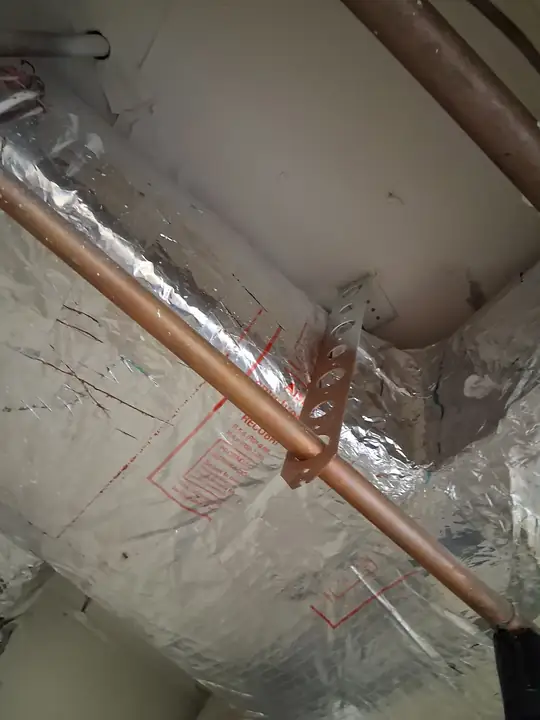 A copper pipe secured with a metal strap beside foil-wrapped insulated ductwork