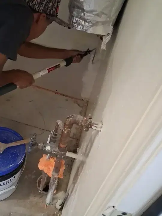 A worker using a long tool on an opened wall above copper supply pipes and valves