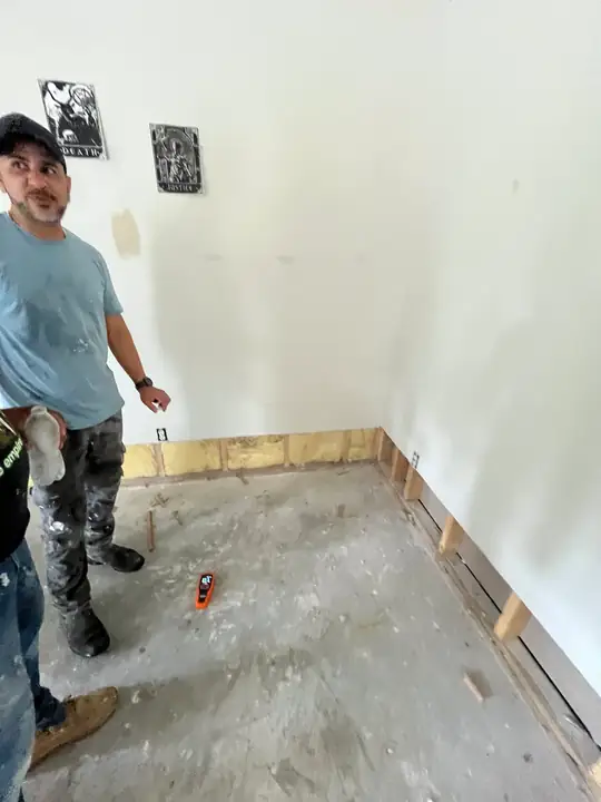 A crew member standing in a room where the bottom of the drywall has been cut away, a moisture meter on the concrete floor
