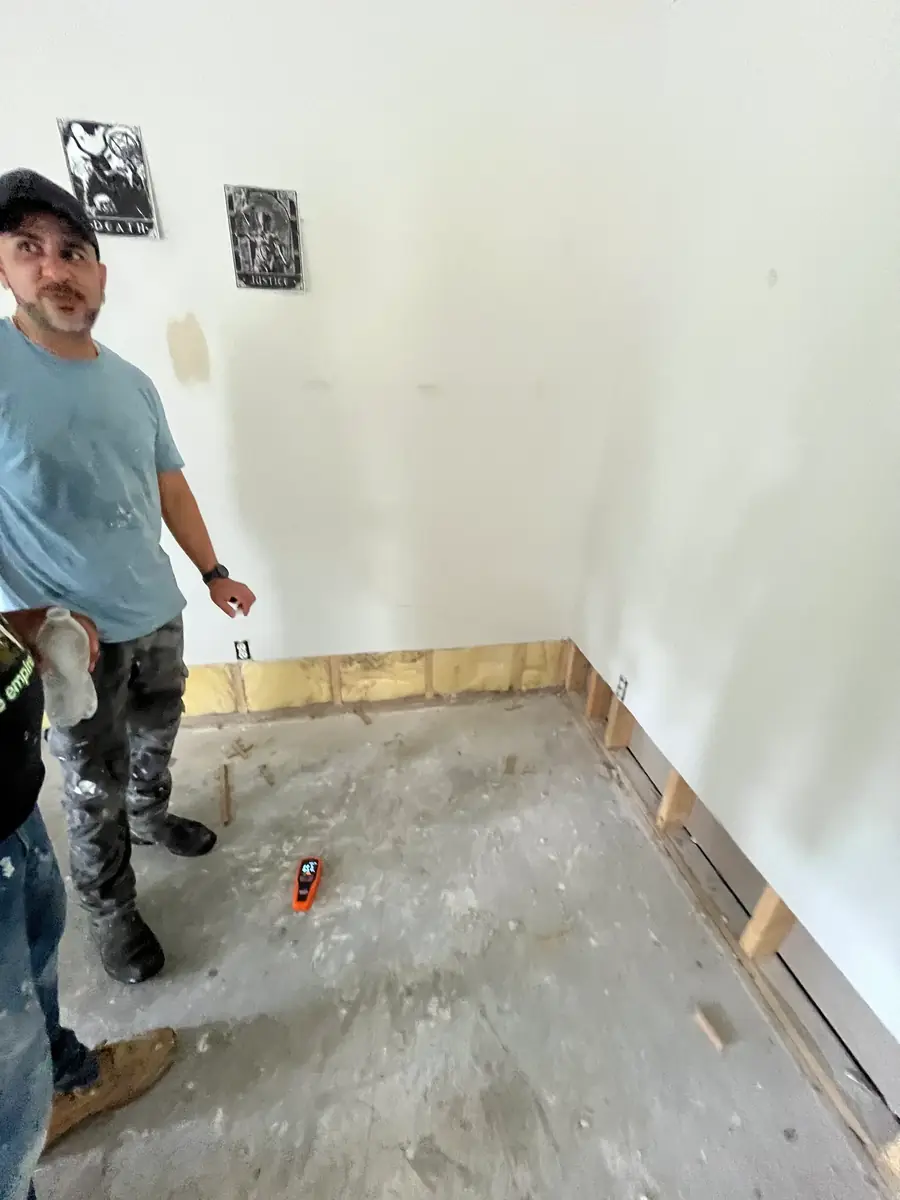 A crew member standing in a room where the bottom of the drywall has been cut away, a moisture meter on the concrete floor