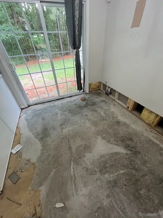 An empty room beside a sliding glass door with the flooring removed to the slab and the lower drywall cut away
