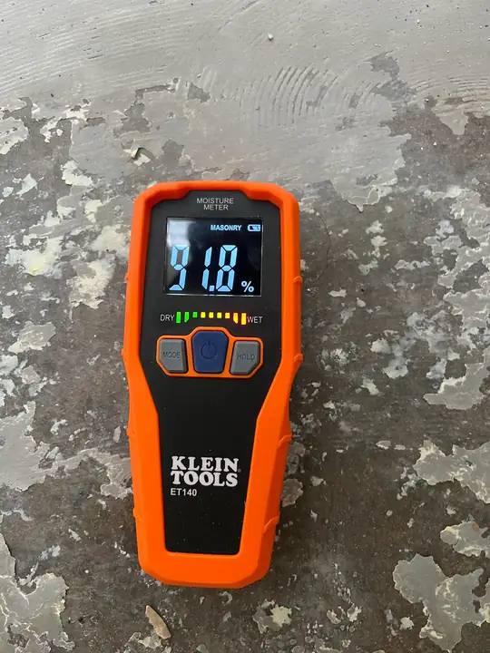 An orange Klein Tools moisture meter reading 91.8 percent, set on a stained concrete slab