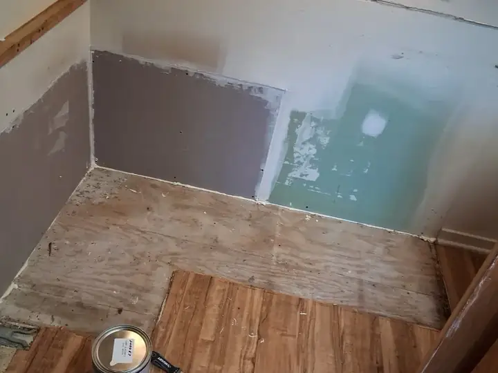 A small room with the flooring pulled back to the plywood subfloor and patched drywall
