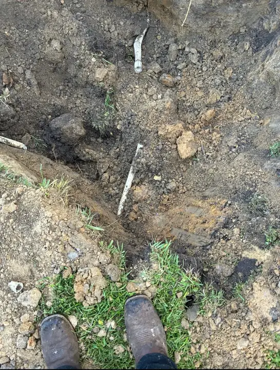 A narrow trench in sandy soil with a white PVC line exposed at the bottom and work boots at the edge